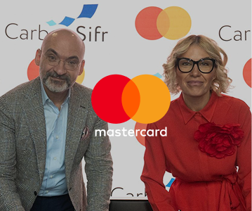 A man in a gray plaid blazer and a woman in a red dress with black glasses posing in front of a backdrop featuring Mastercard and CarboSifr logos.