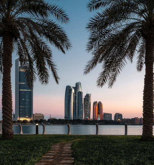 City skyline with modern skyscrapers at sunset, viewed through two palm trees by a waterfront park.