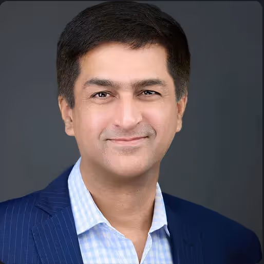 Headshot of a man with short black hair wearing a blue pinstripe suit jacket and light blue checkered shirt, smiling gently against a dark background.