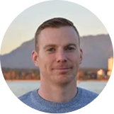 Portrait of a man with short light brown hair wearing a blue shirt, with mountains and buildings in the background.
