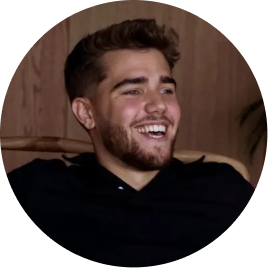 Young man with short hair and beard smiling and looking to the side, seated indoors with a wooden background.