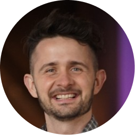 Smiling man with short curly hair and a trimmed beard in a circular frame.