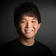 Portrait of a smiling young man with short dark hair wearing a black shirt against a dark background.