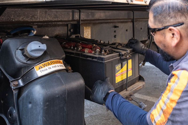 Mechanic wearing gloves and safety glasses installs or inspects a large truck battery mounted under the chassis near fuel tank.