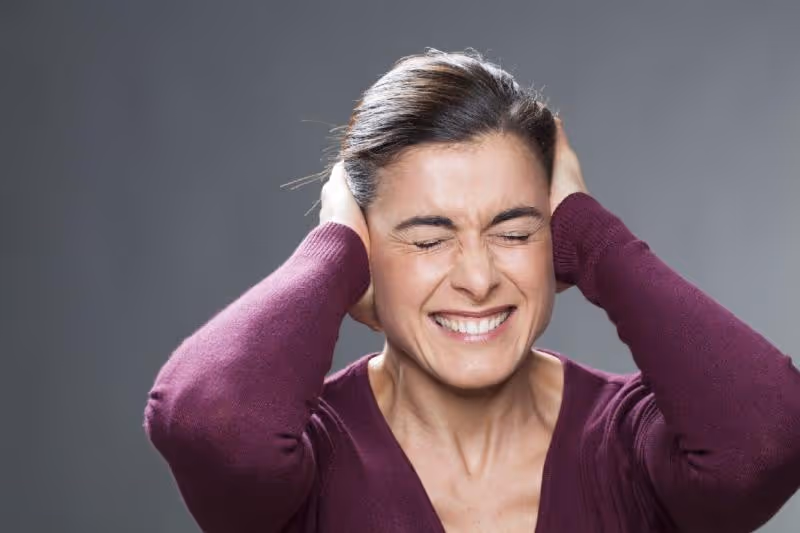 A woman clenching her teeth with closed eyes and holding her hands over her ears