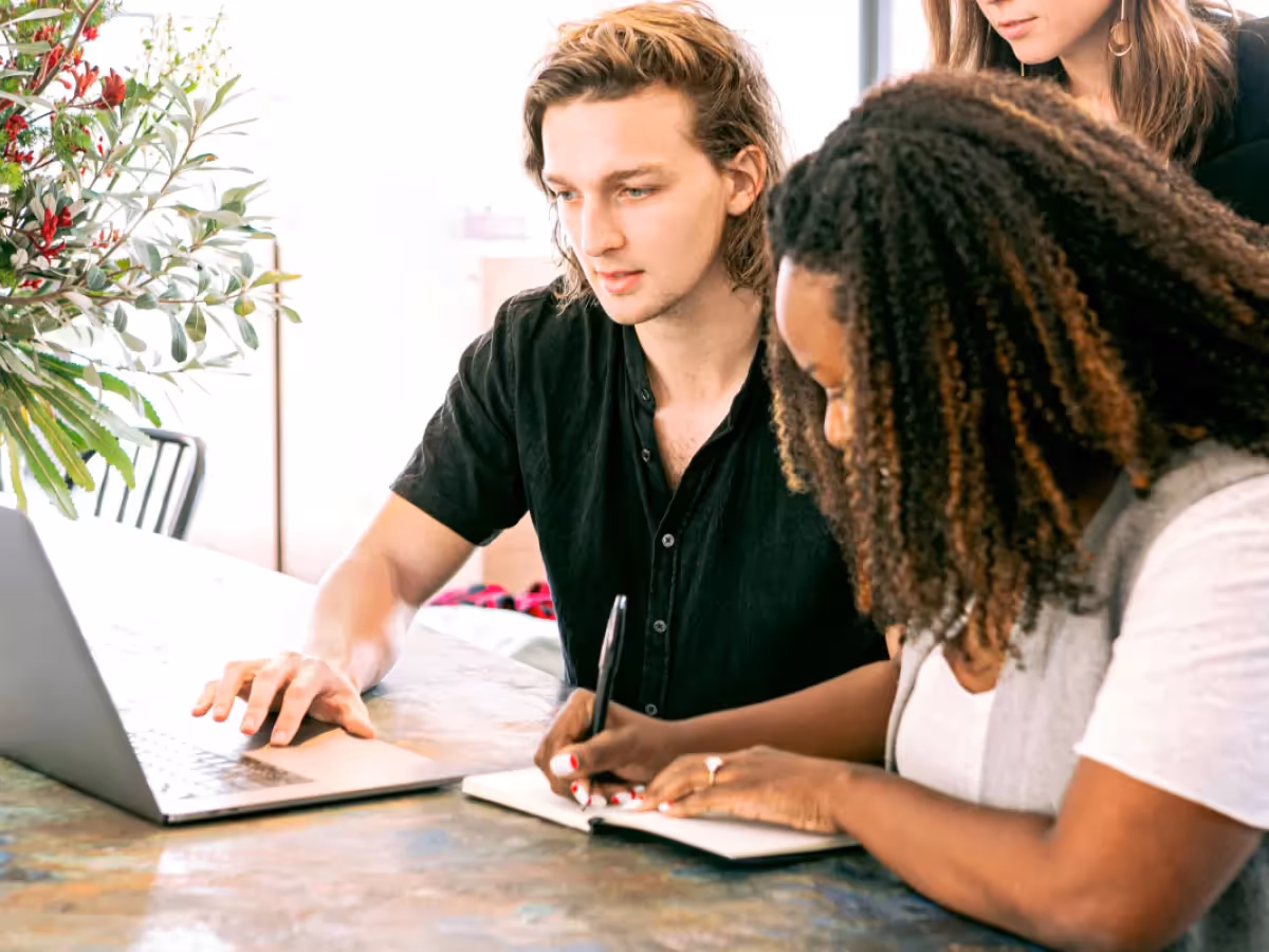 A collaborative team working at a table, featuring a young man using a laptop and guiding a woman taking notes in a notebook, with another person standing behind them
