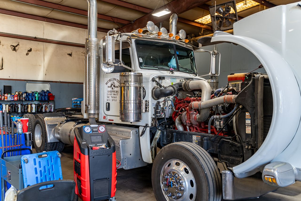 Semi-truck with hood open inside repair shop, showing exposed engine and diagnostic tools, surrounded by parts, fluids, and equipment. 