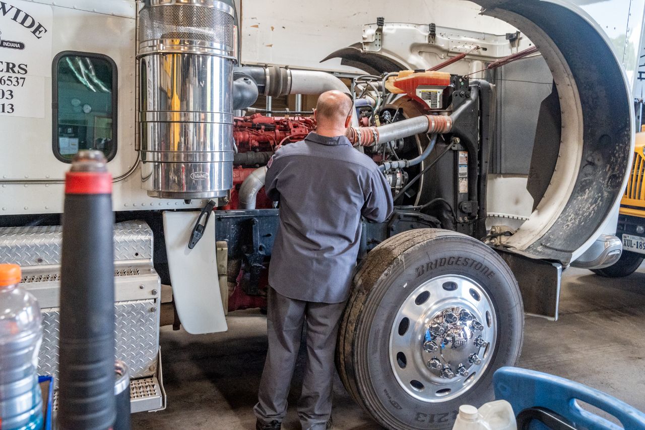 Mechanic inspecting and repairing a semi-truck engine with the hood open inside a workshop, focusing on engine components and connections.