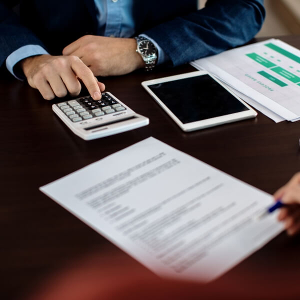 Une personne en costume utilisant une calculatrice sur un bureau avec des documents financiers et une tablette.