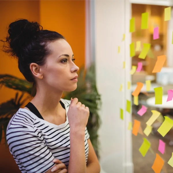 Une femme observe un mur recouvert de notes autocollantes colorées, réfléchissant à des idées ou des projets.