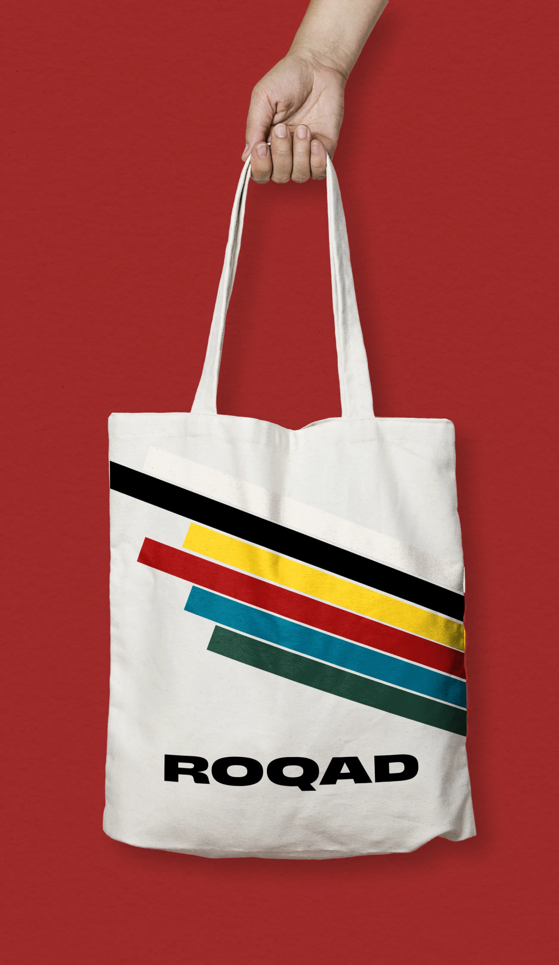 White tote bag with diagonal stripes in black, yellow, red, blue, and green, and the word ROQAD printed below the stripes, held by a hand against a red background.