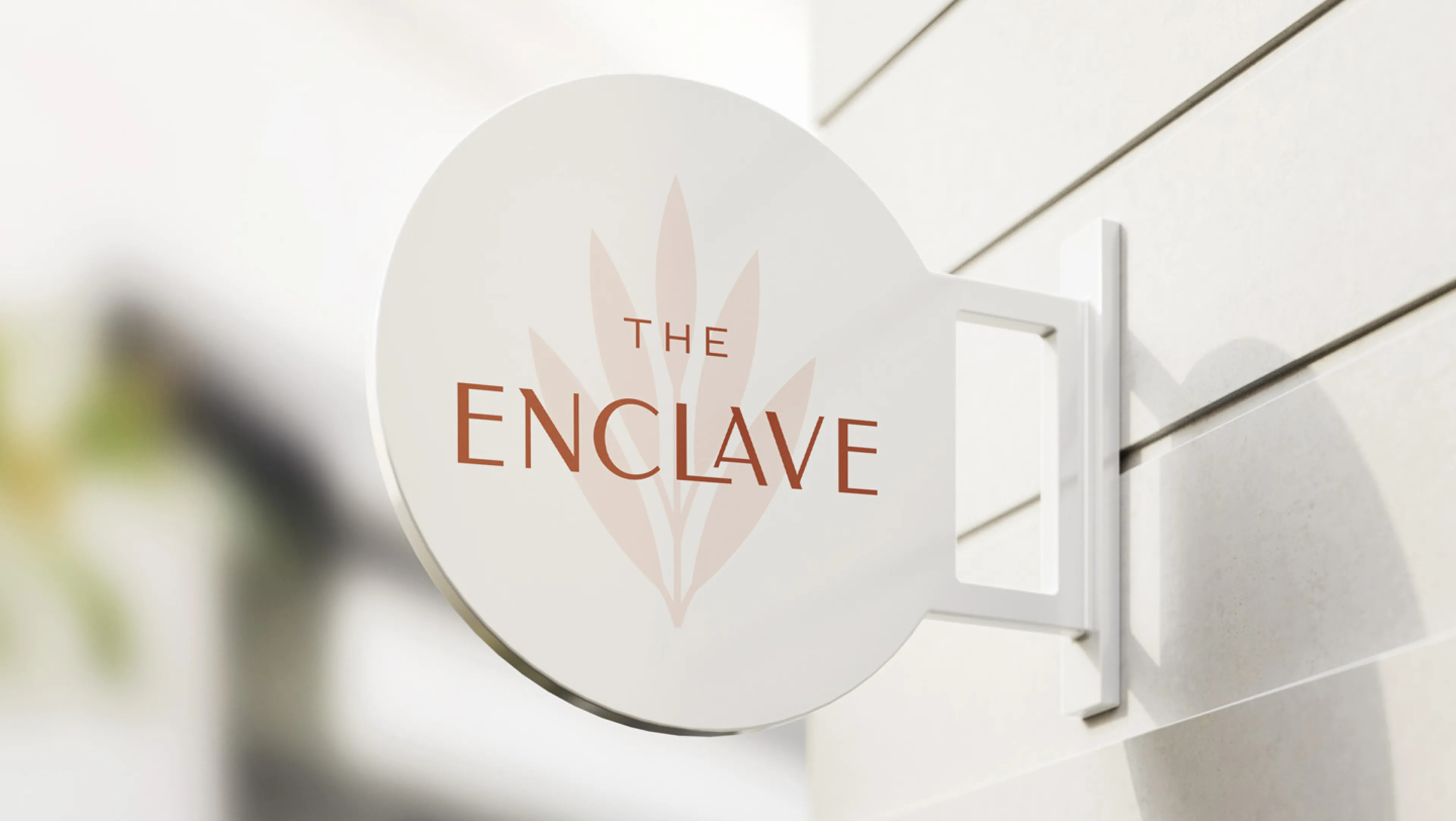 Round white wall-mounted sign with a subtle leaf design and the text 'THE ENCLAVE' in brown.