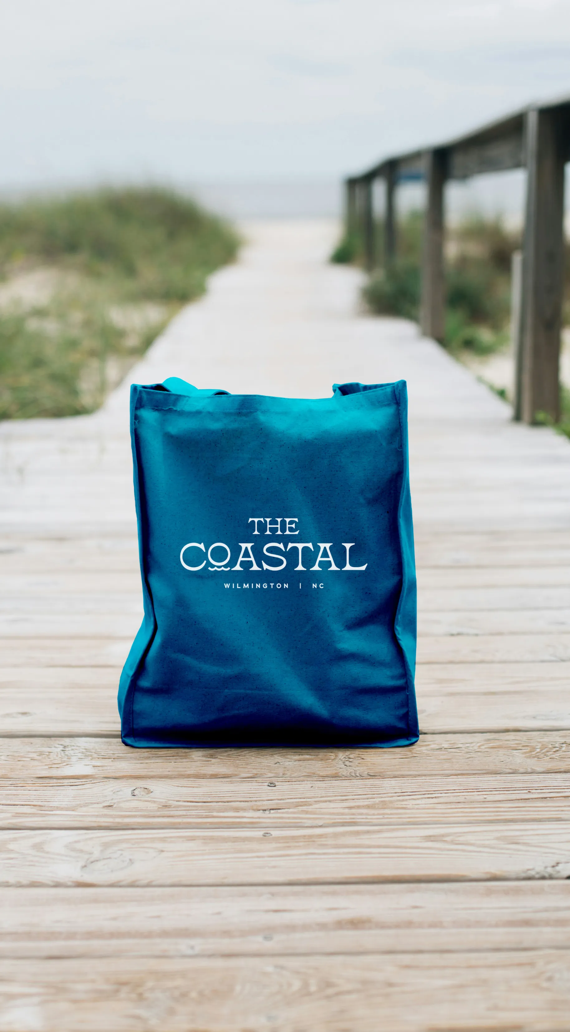 Blue tote bag with 'The Coastal Wilmington NC' text, placed on a wooden boardwalk near beach dunes.