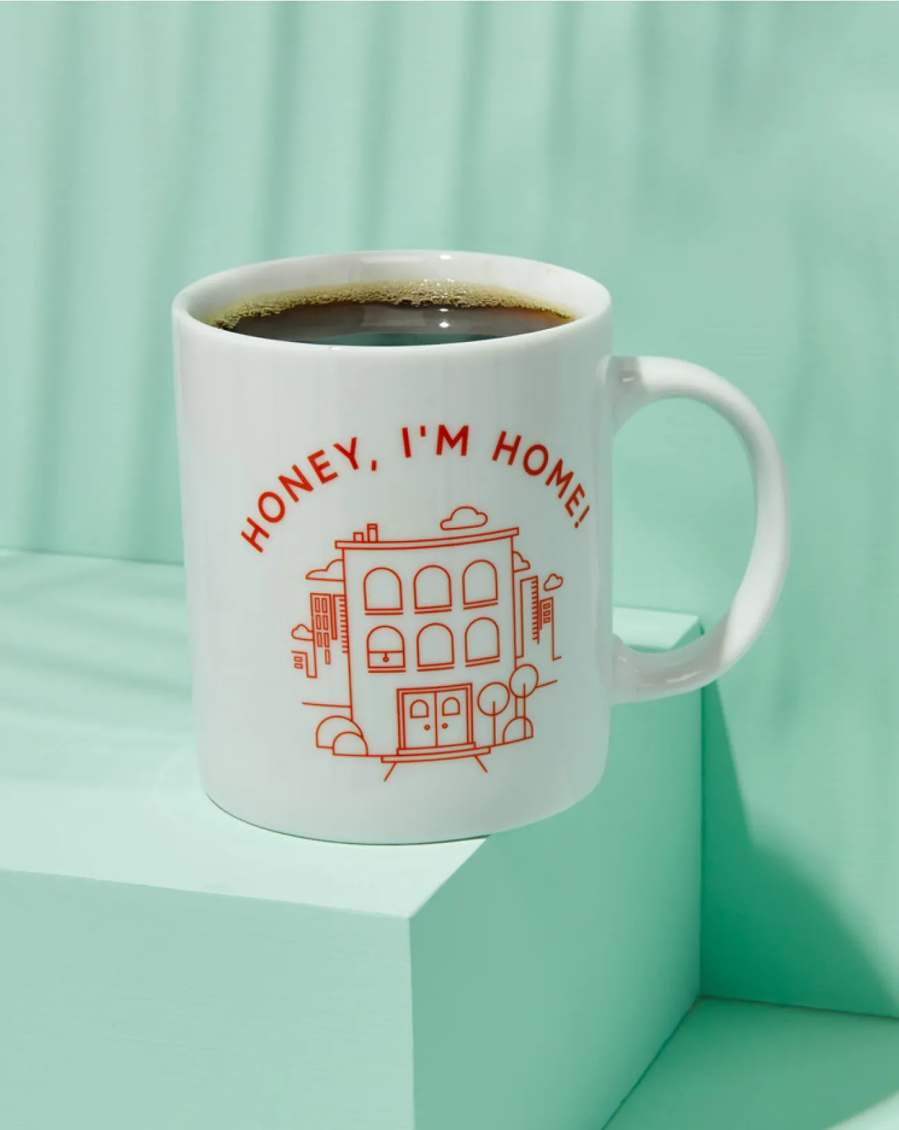 White coffee mug with red text 'HONEY, I'M HOME!' and a line drawing of a house, filled with black coffee, on a light teal block and background.