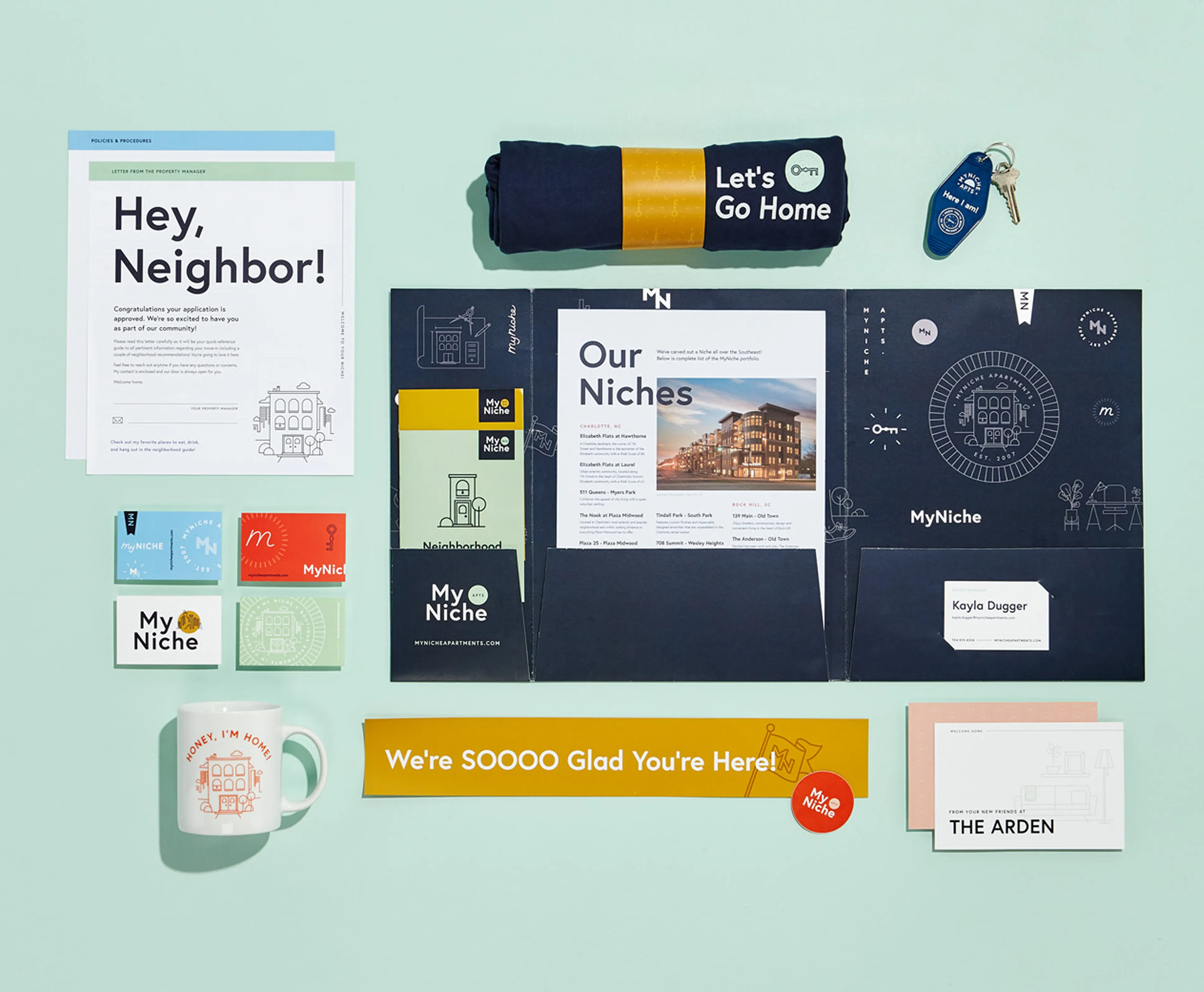 Flat lay of MyNiche branded welcome kit including a folder with brochures, letter, business card, keys with keychain, mug, t-shirt, stickers, and welcome cards on a light blue background.