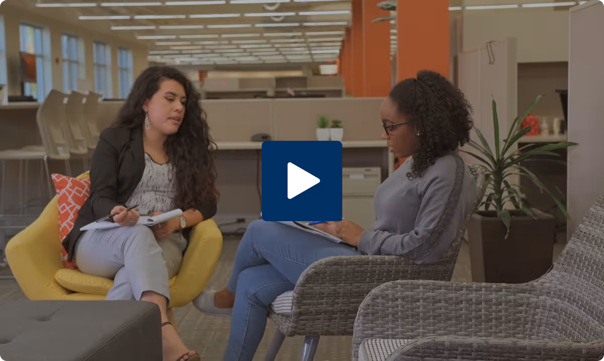 Two women seated and having a discussion in a modern office lounge area with note-taking materials.
