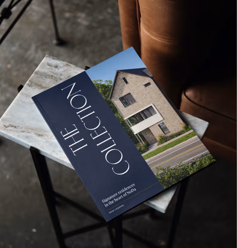 A brochure titled 'The Collection' featuring a modern brick house, placed on a marble table near a brown leather chair.