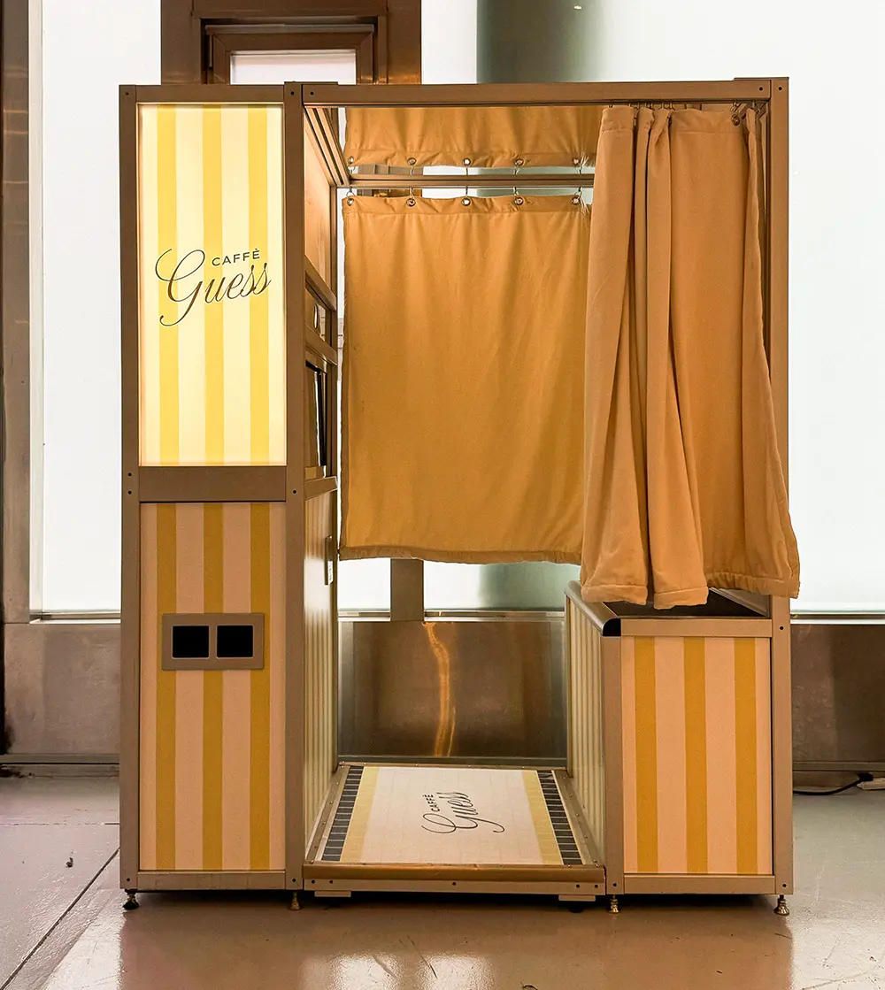 Front view of the Caffè Guess cafe photo booth showing striped paneling, curtain, and branded floor mat