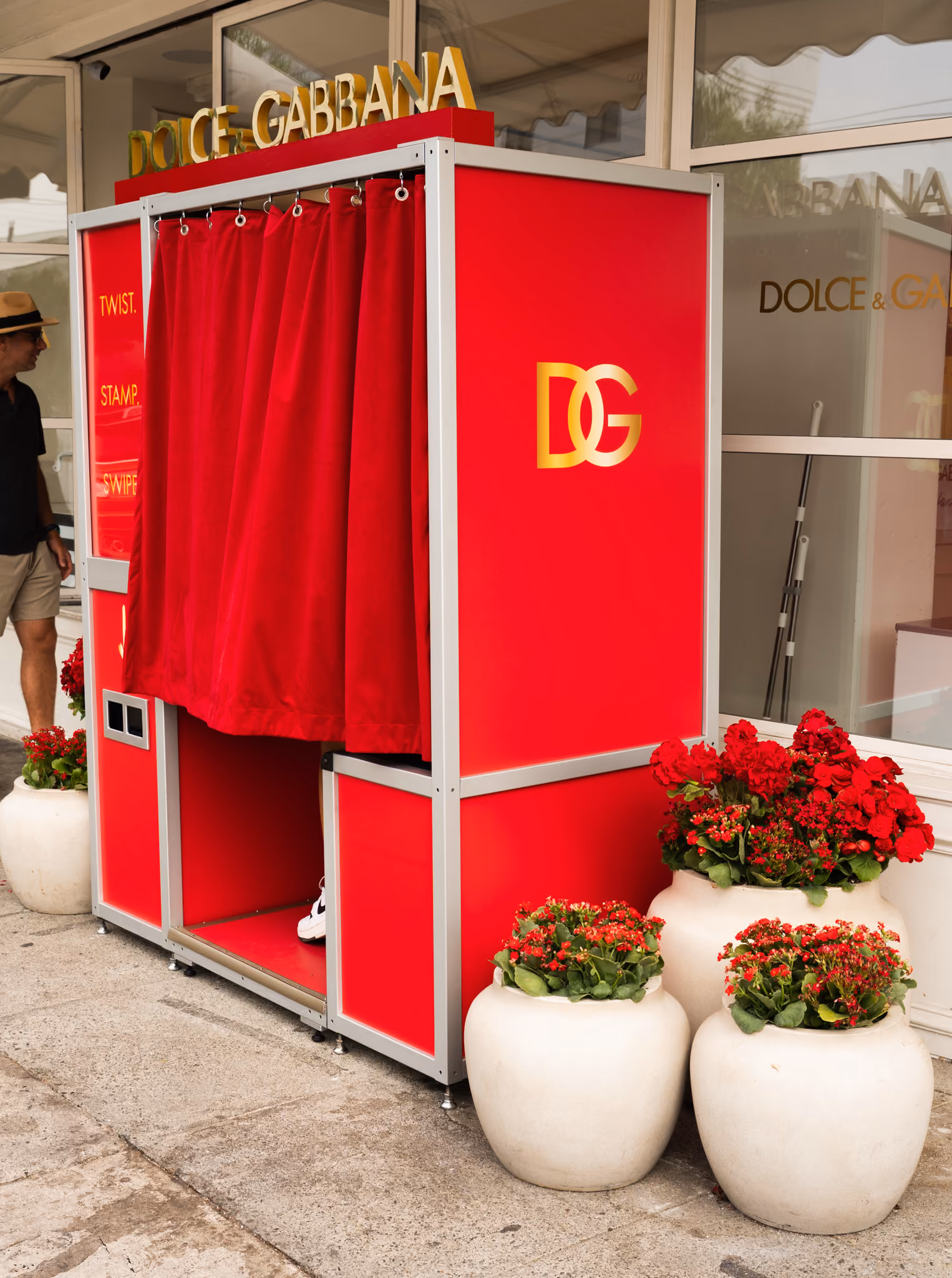 Red Dolce & Gabbana photo booth with a gold DG monogram on the side panel, set on the pavement beside red flowering planters
