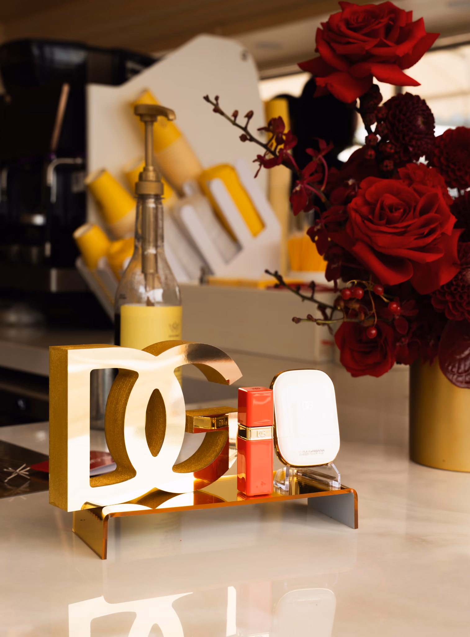 Gold DG monogram block on the cafe counter beside a red Dolce & Gabbana lipstick and compact, with dark red roses behind