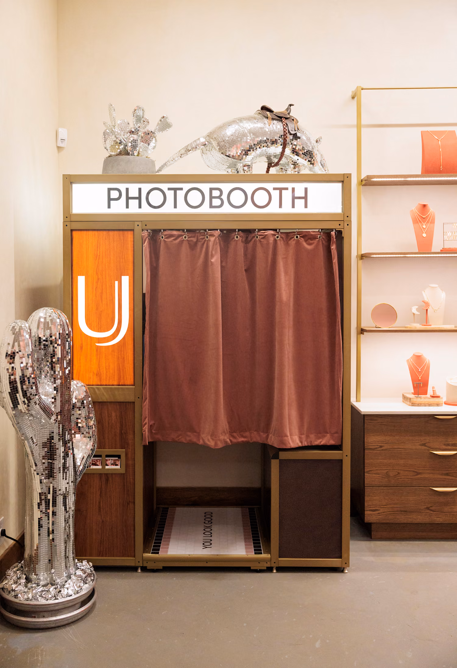 Gold framed photo booth with an illuminated PHOTOBOOTH marquee and a mauve velvet curtain inside the Uncommon James store in Austin, with mirrored disco cactus sculptures beside it