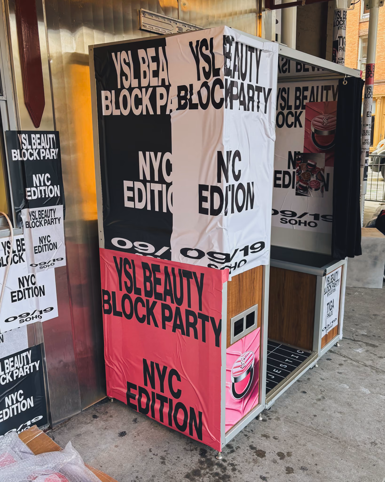 Photo booth postered in pink and black YSL Beauty Block Party artwork on a Soho sidewalk, with matching flyposters on the wall behind it