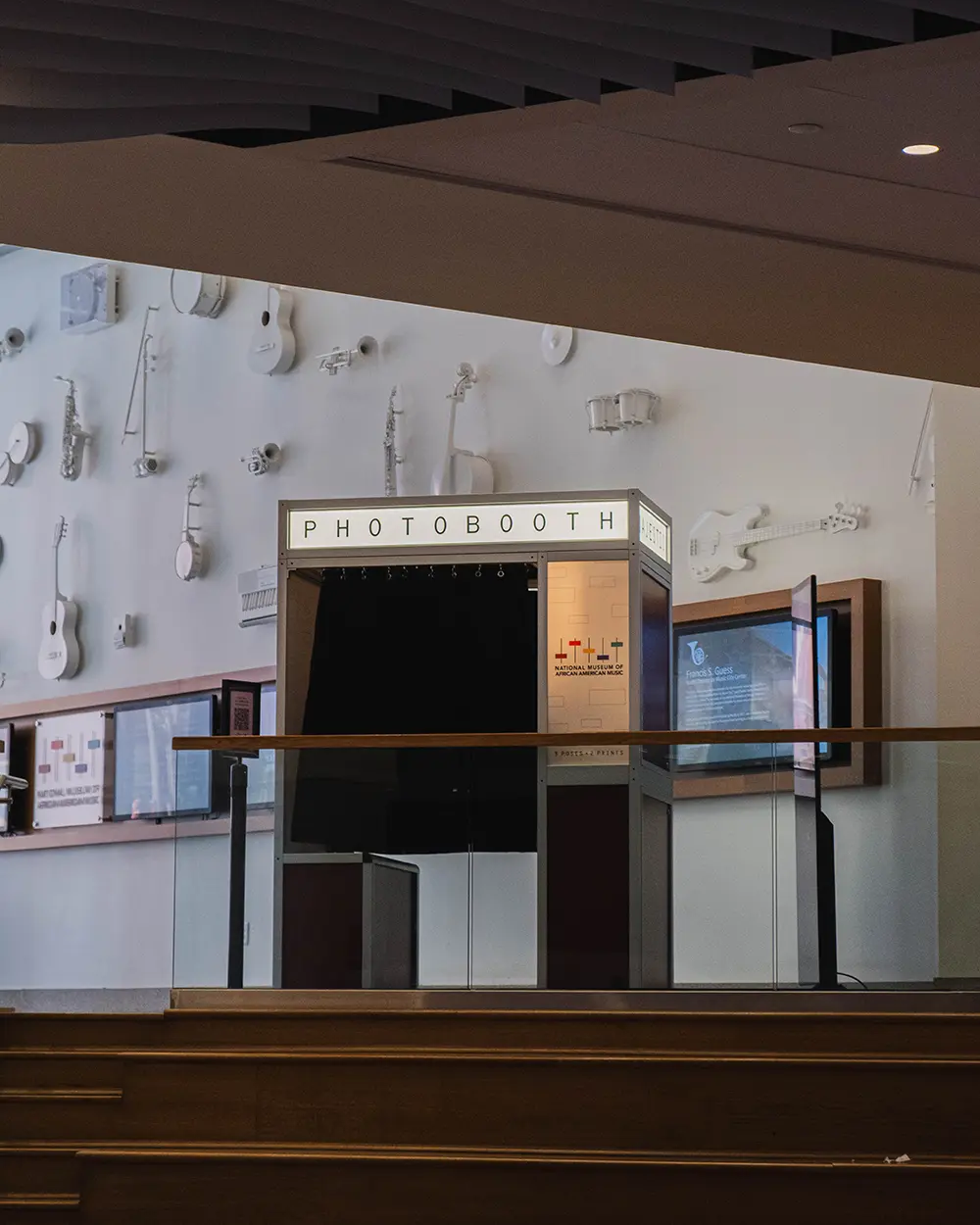 Photo booth with a lit PHOTOBOOTH marquee installed on the Grand Foyer landing at the National Museum of African American Music, below a wall of white sculptural instruments