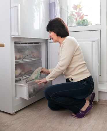 Woman kneeling on the floor in front of an open freezer drawer, placing or removing a sealed plastic bag of food.