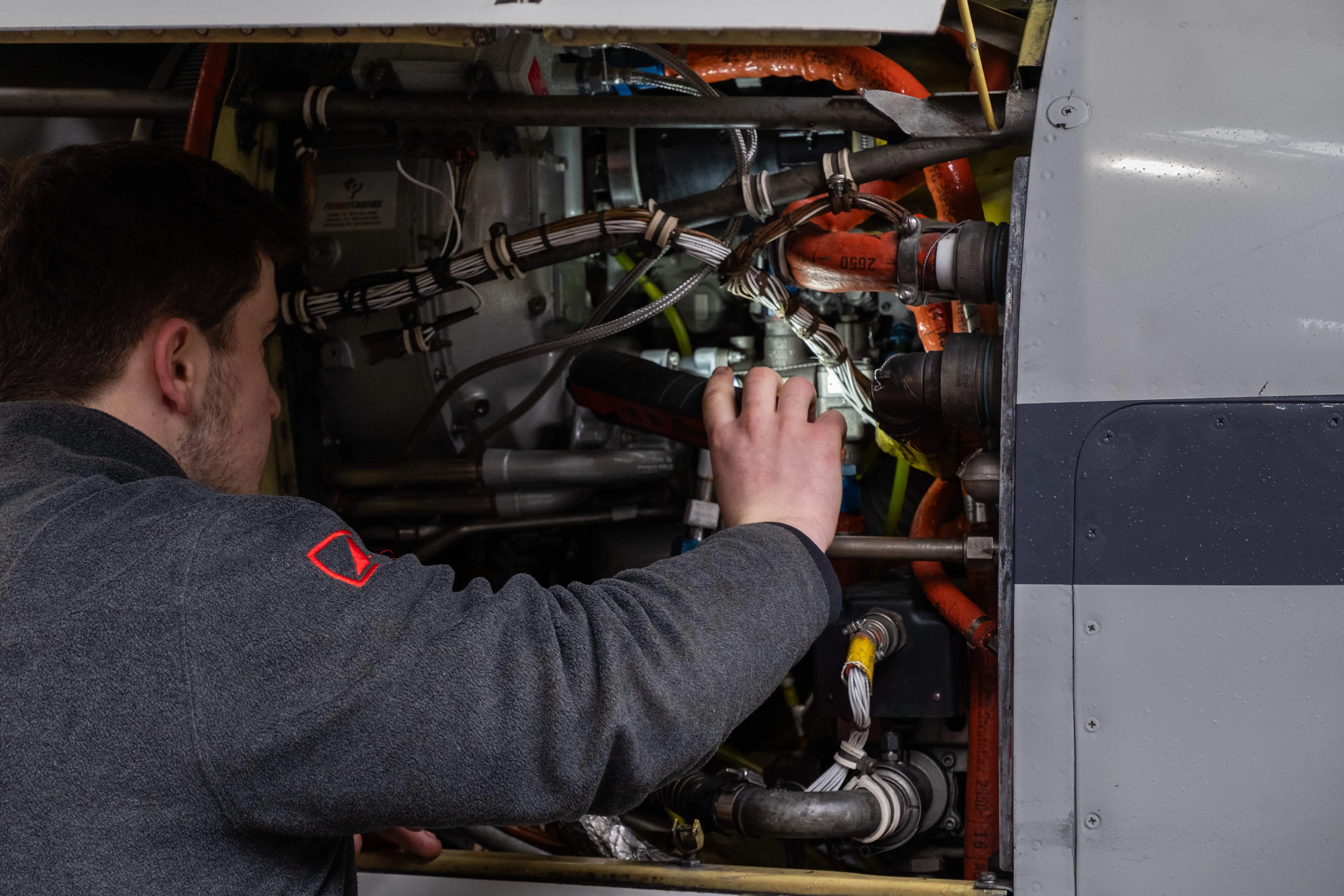 Un homme examine et travaille sur un moteur complexe avec plusieurs câbles, tuyaux et composants métalliques visibles.