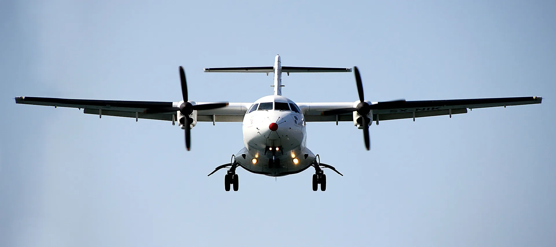 Avion turbopropulseur blanc vu de face en vol avec les trains d'atterrissage sortis et les hélices en rotation sur un ciel bleu clair.