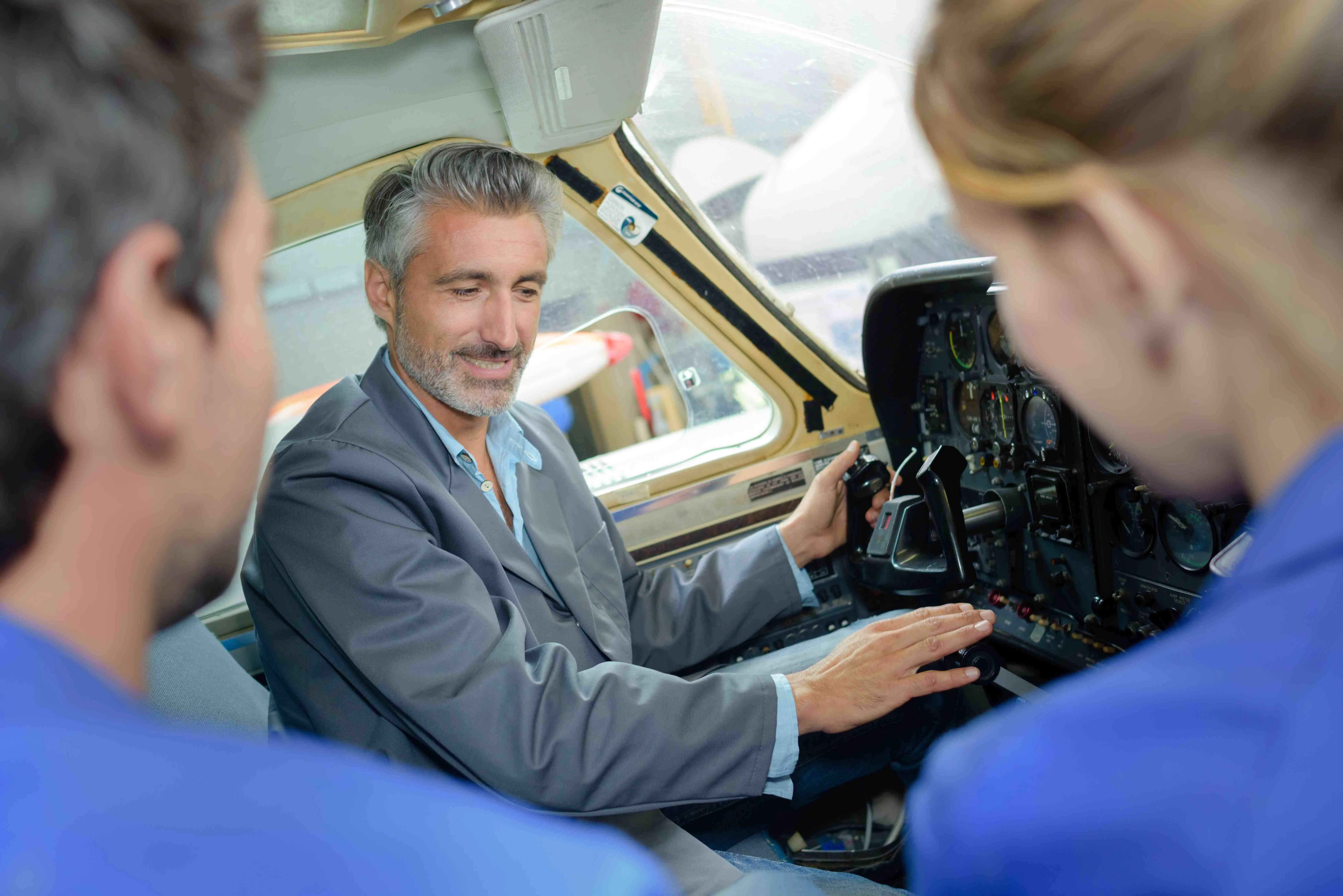 Un homme souriant, habillé d'une veste grise, montre les commandes dans le cockpit d’un petit avion à deux personnes habillées en bleu, observant attentivement.