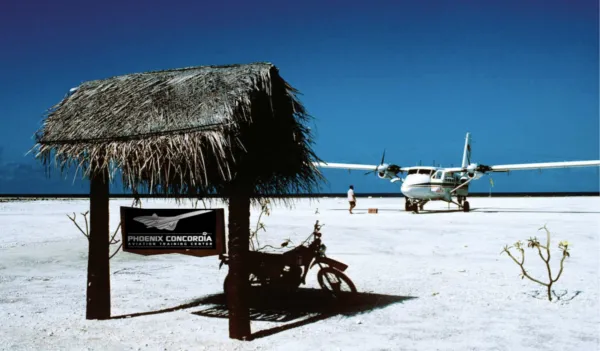 Petit avion bimoteur blanc stationné sur une grande étendue de surface blanche, avec un homme debout près de l'appareil volant, et un abri en bois avec toit en feuilles sèches et une moto garée à l'ombre, panneau indiquant Phoenix Concordia Aviation Training Centre.