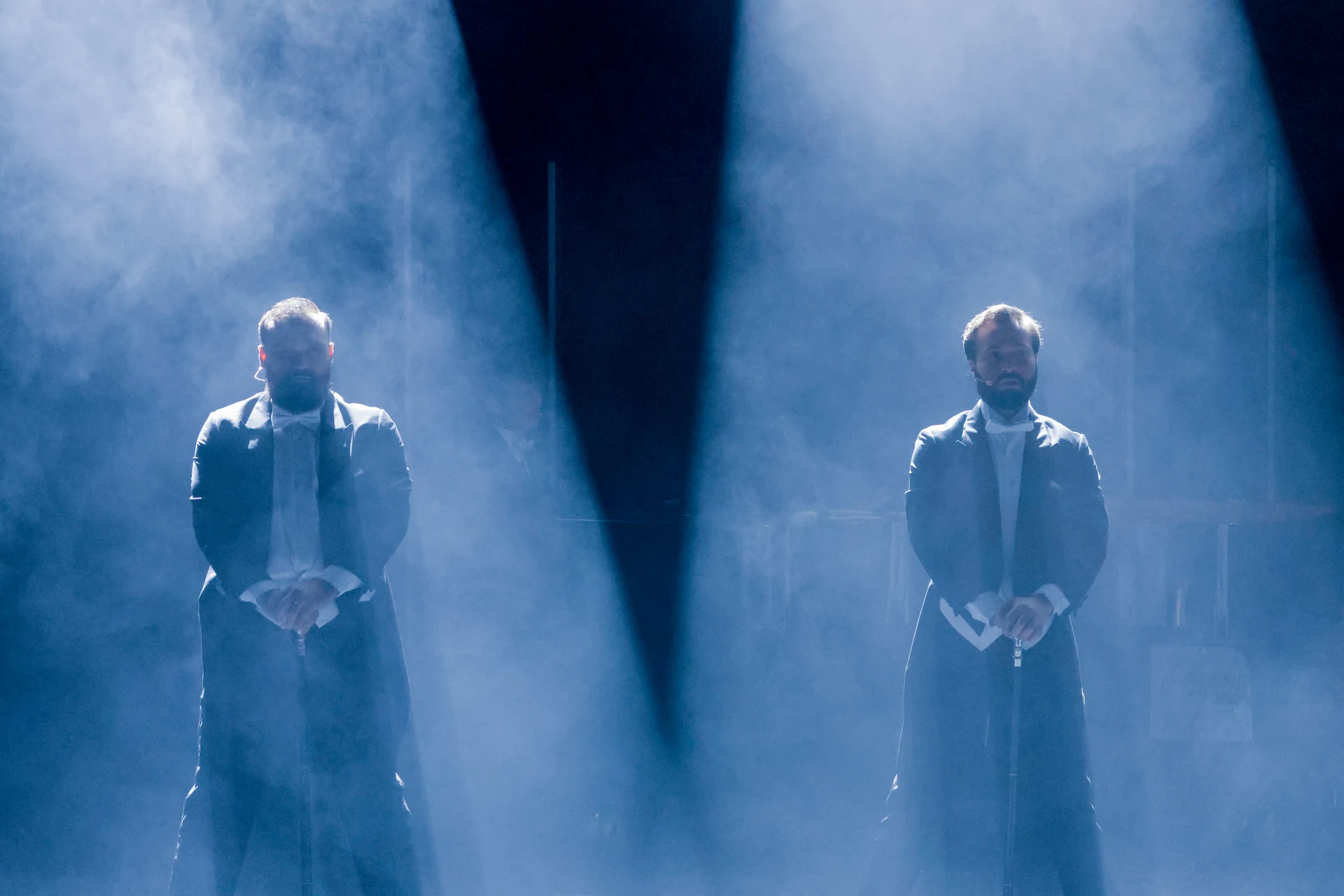 Twee zangers staan onder blauwe en witte schijnwerpers in theaternevel.