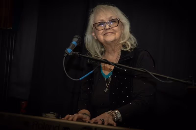 Smiling elderly woman with glasses playing keyboard in front of a microphone on a dark background.