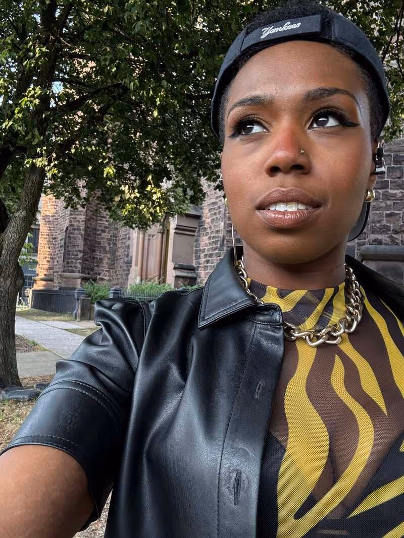 Person wearing a black Yankees cap, black leather jacket, gold chain necklace, and a yellow and brown patterned top, standing outdoors near a tree and stone building.