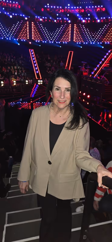 Woman with dark hair wearing a beige blazer and black top standing on stairs in a concert venue with bright stage lights and audience in the background.