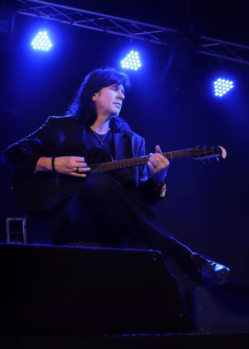 Man playing an acoustic guitar on stage under blue spotlights, dressed in black with long dark hair.