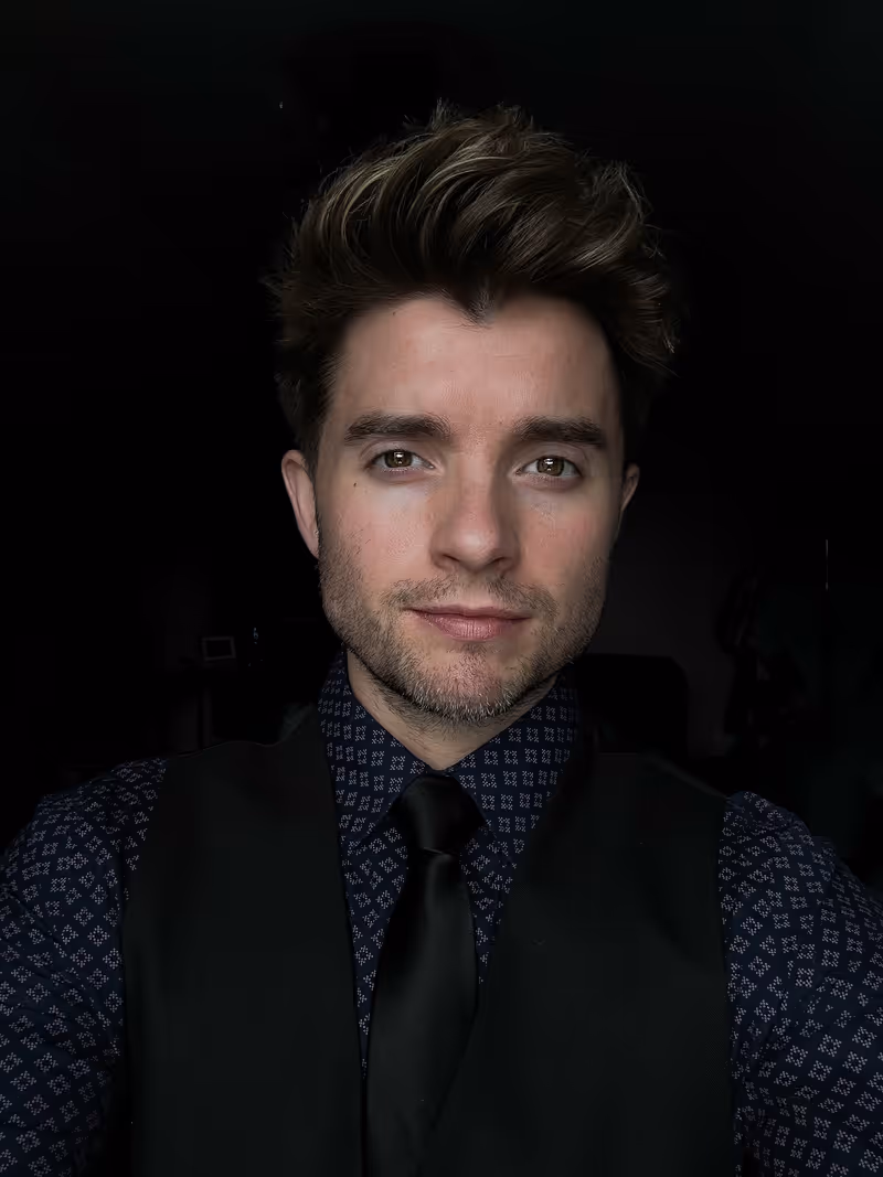 Portrait of a young man with styled hair wearing a patterned shirt, black vest, and black tie against a dark background.