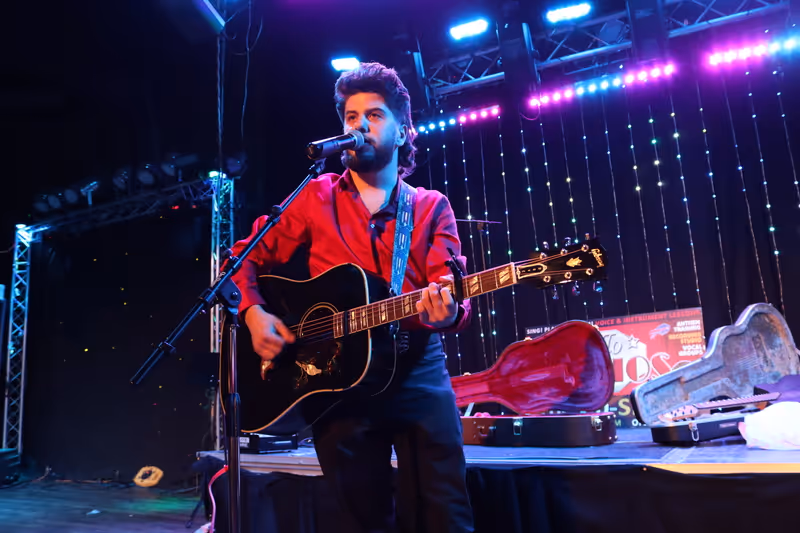 Musician playing black acoustic guitar and singing into microphone on stage with colorful lights in the background.