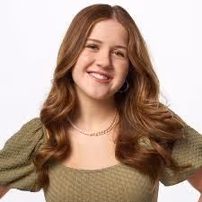 Smiling young woman with long wavy brown hair wearing a textured green top and a necklace.
