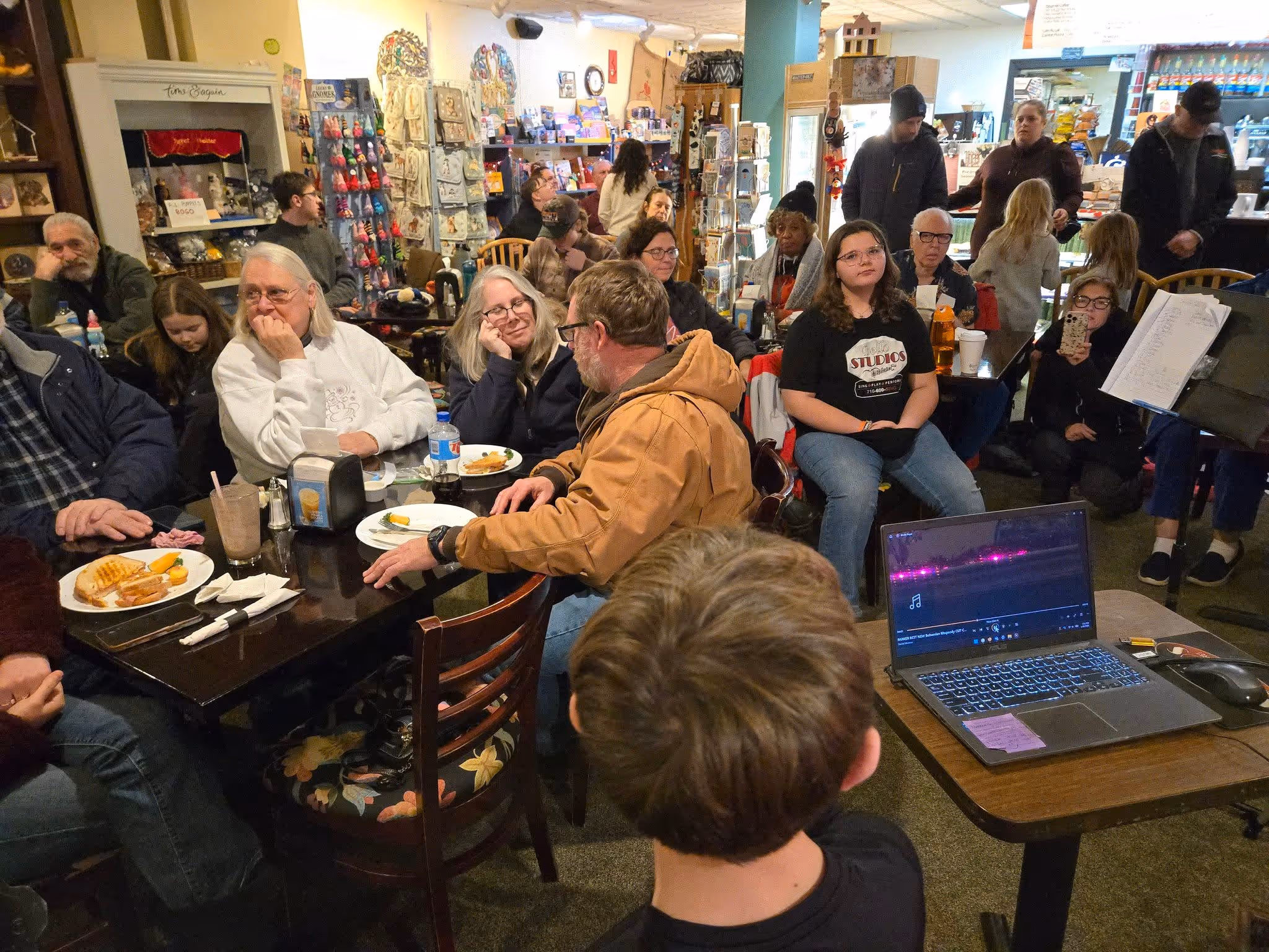 Audience of diverse ages seated and standing in a cozy café watching a person presenting with a laptop.