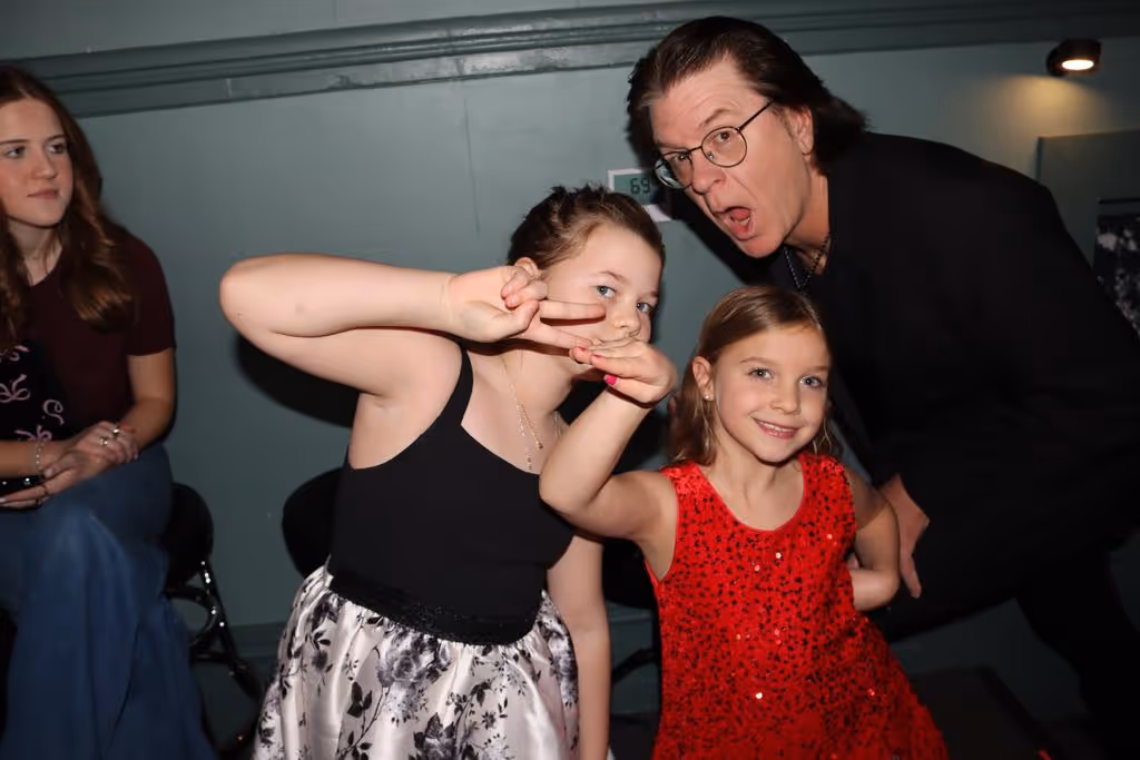 Two young girls smiling and posing with peace signs while a man with glasses leans in from behind making a funny face, with a woman sitting in the background.