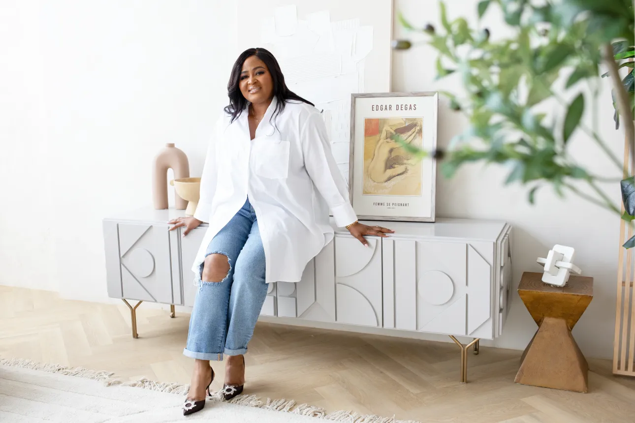 Woman in white shirt and ripped jeans sitting on a modern white sideboard with decorative objects and an Edgar Degas artwork behind her.