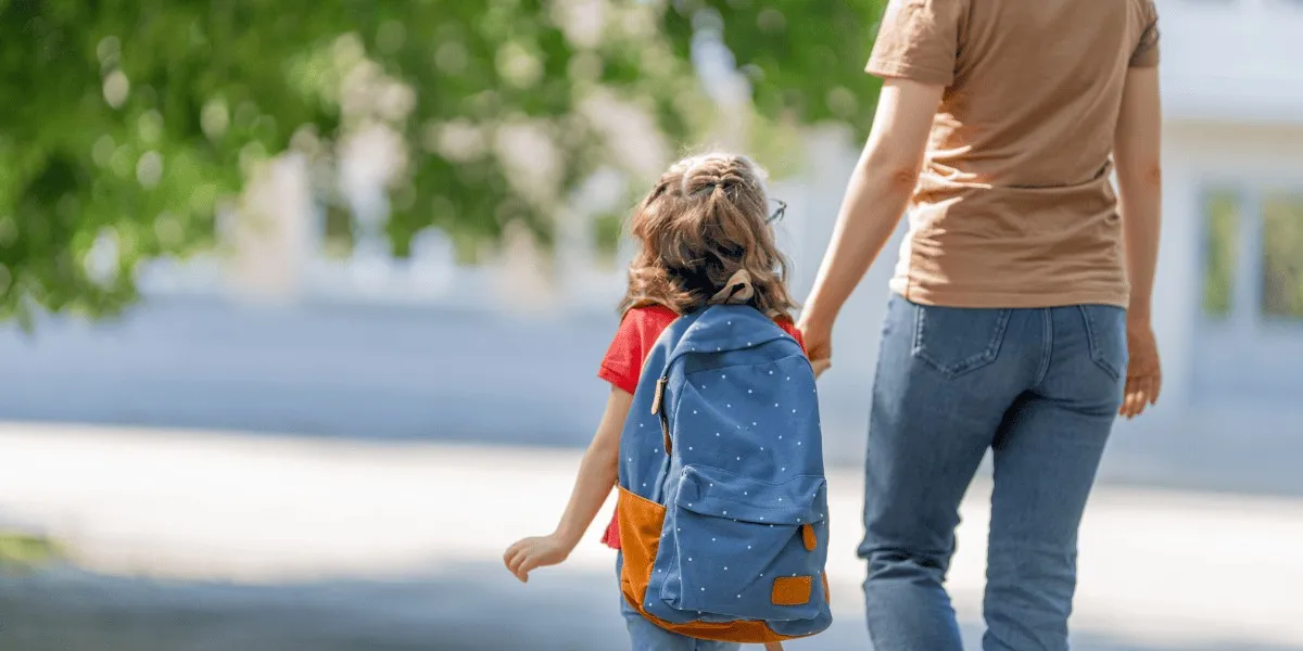 A woman and a young girl with a blue backpack are walking together, holding hands, with green trees and a building in the blurred background.