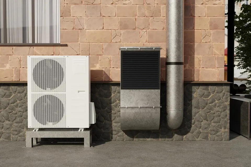 A new white outdoor AC unit is placed on a metal stand next to a gray metal ventilation pipe and a black metal box, with a pink brick wall and gray stone siding behind it.