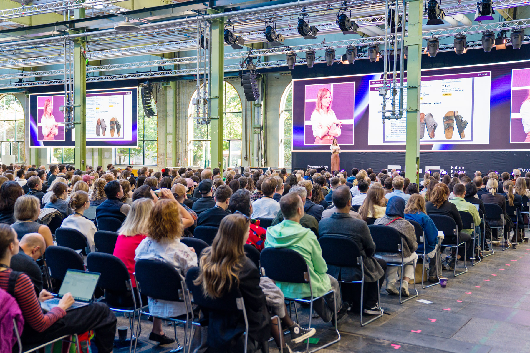 Large audience seated indoors watching a woman giving a presentation on stage with her image and slides displayed on large screens.