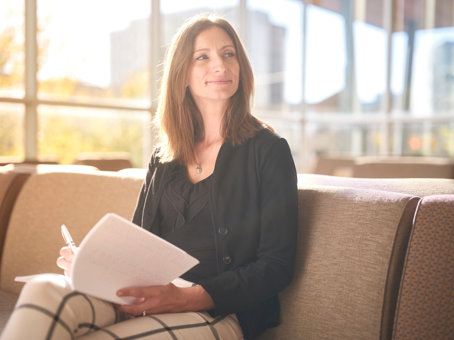 A alumni smiling, sitting on a couch with the sun shining behind her.
