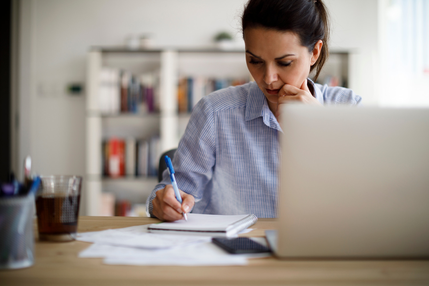 A woman writing in her notebook with a laptop in front of her.