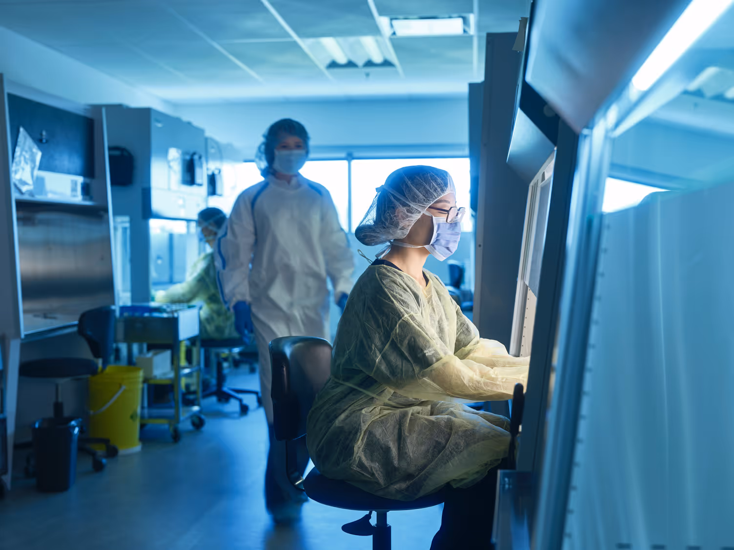 Pharmacy technician students in protective gear and face masks work with equipment and computers in a brightly lit lab, focusing on analysis and research. One person sits at a workstation while others move in the background.