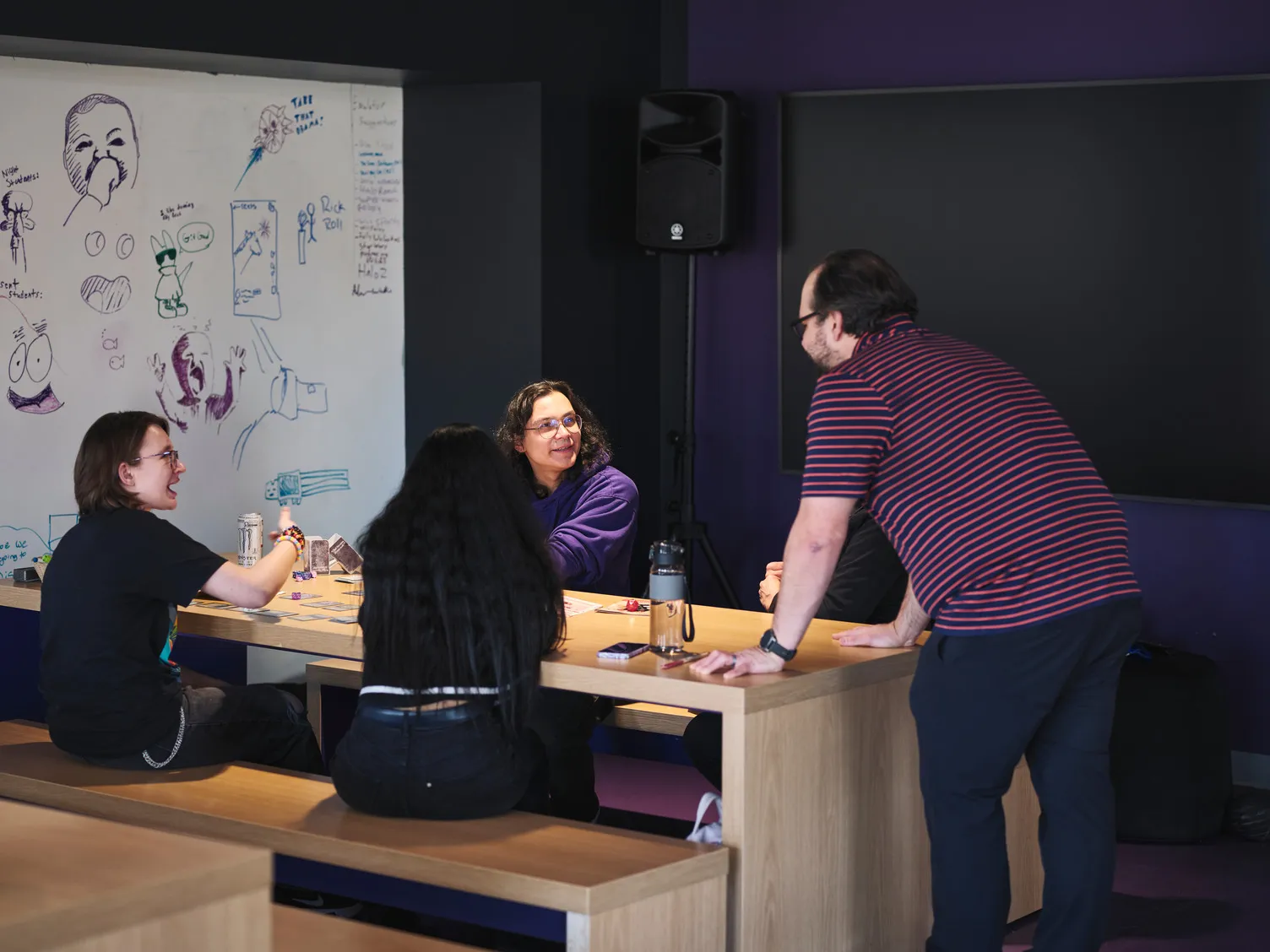 Four people sit and talk around a wooden table in a room with purple walls. A whiteboard behind them is covered in doodles, cartoon faces, and sketches. One person stands, engaging with the others, who are smiling.